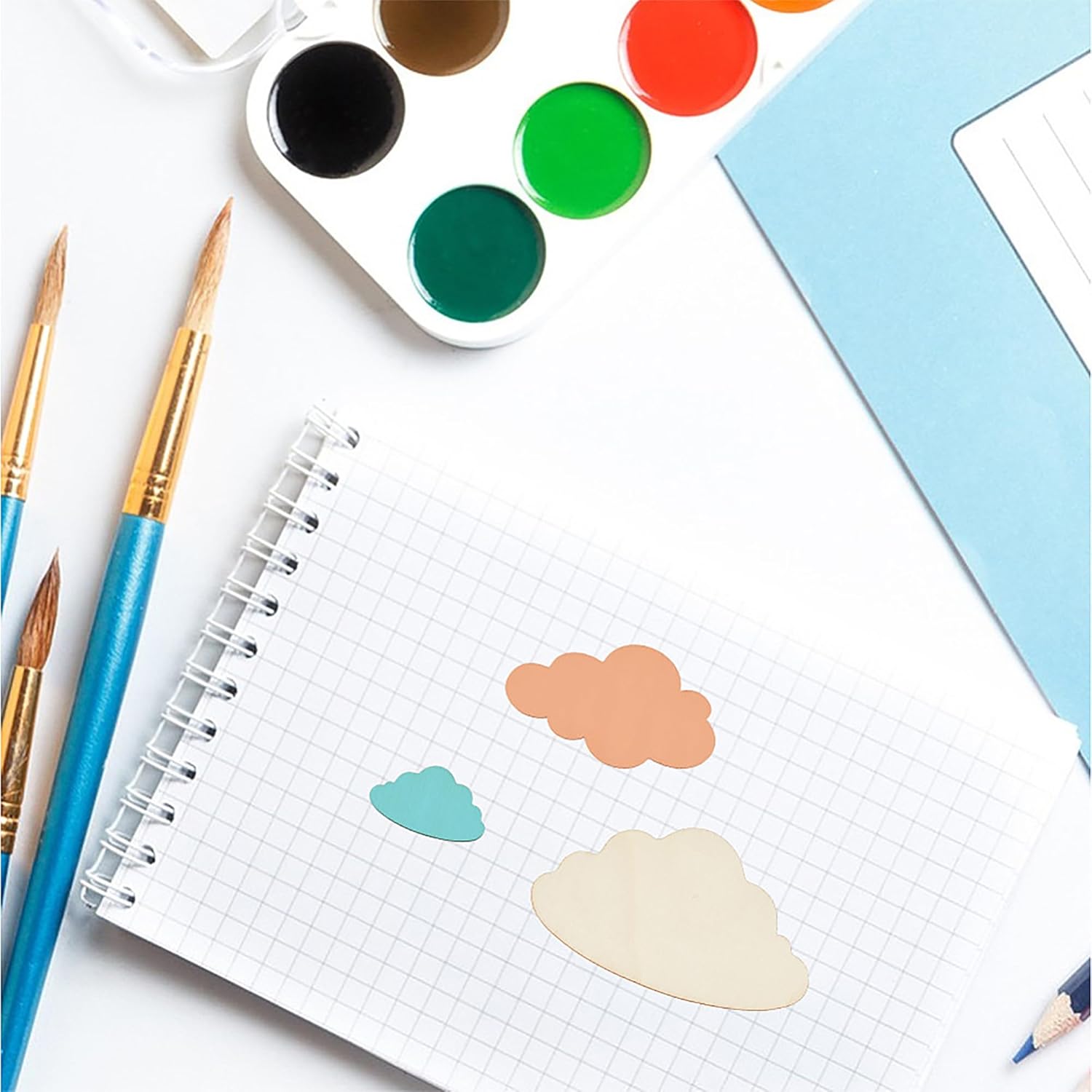Art supplies including watercolor paints, brushes, and cloud-shaped stickers on a notebook.
