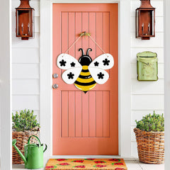 Pink door with a bee-shaped decoration, green watering can, and planters on a white wall.