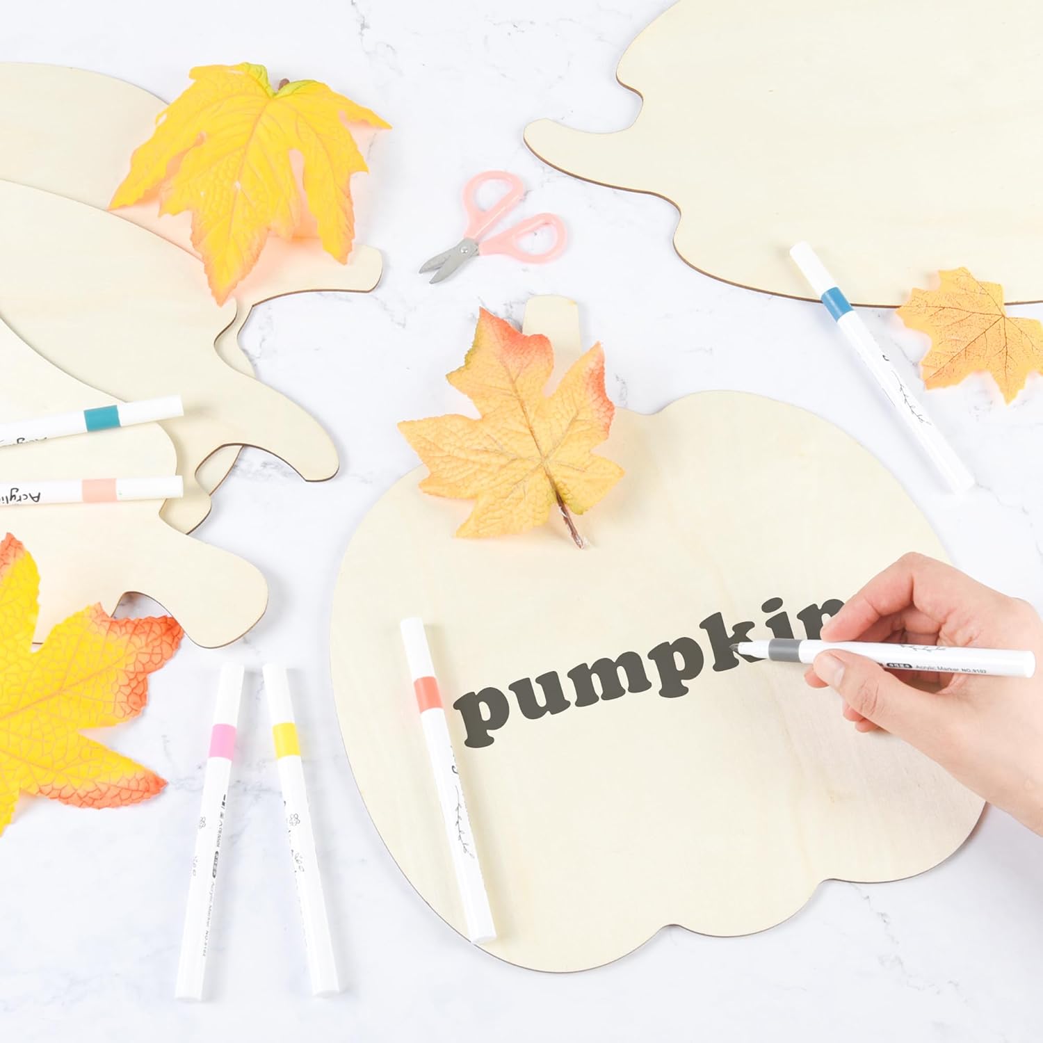 Handwriting 'pumpkin' on a leaf-shaped card with autumn leaves and markers on a white background