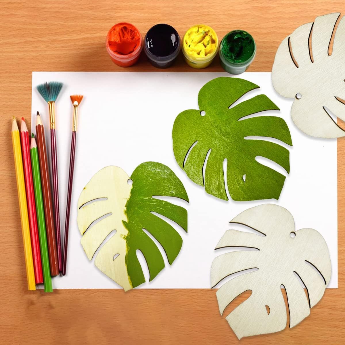 Art supplies including paint, brushes, and colored pencils on a wooden surface with leaf-shaped cutouts.