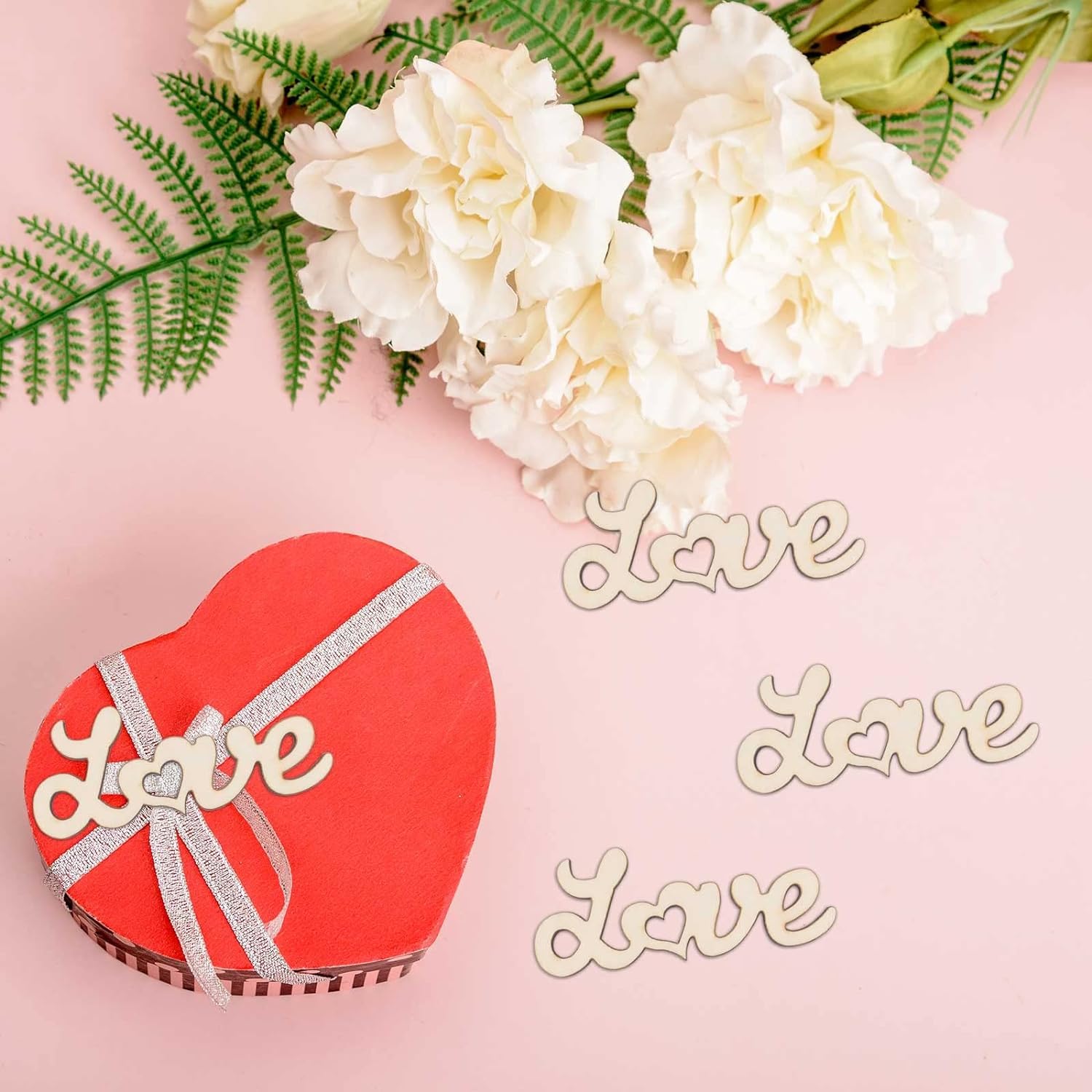 Heart-shaped red gift box with 'love' text, surrounded by white flowers and green leaves on a pink background.