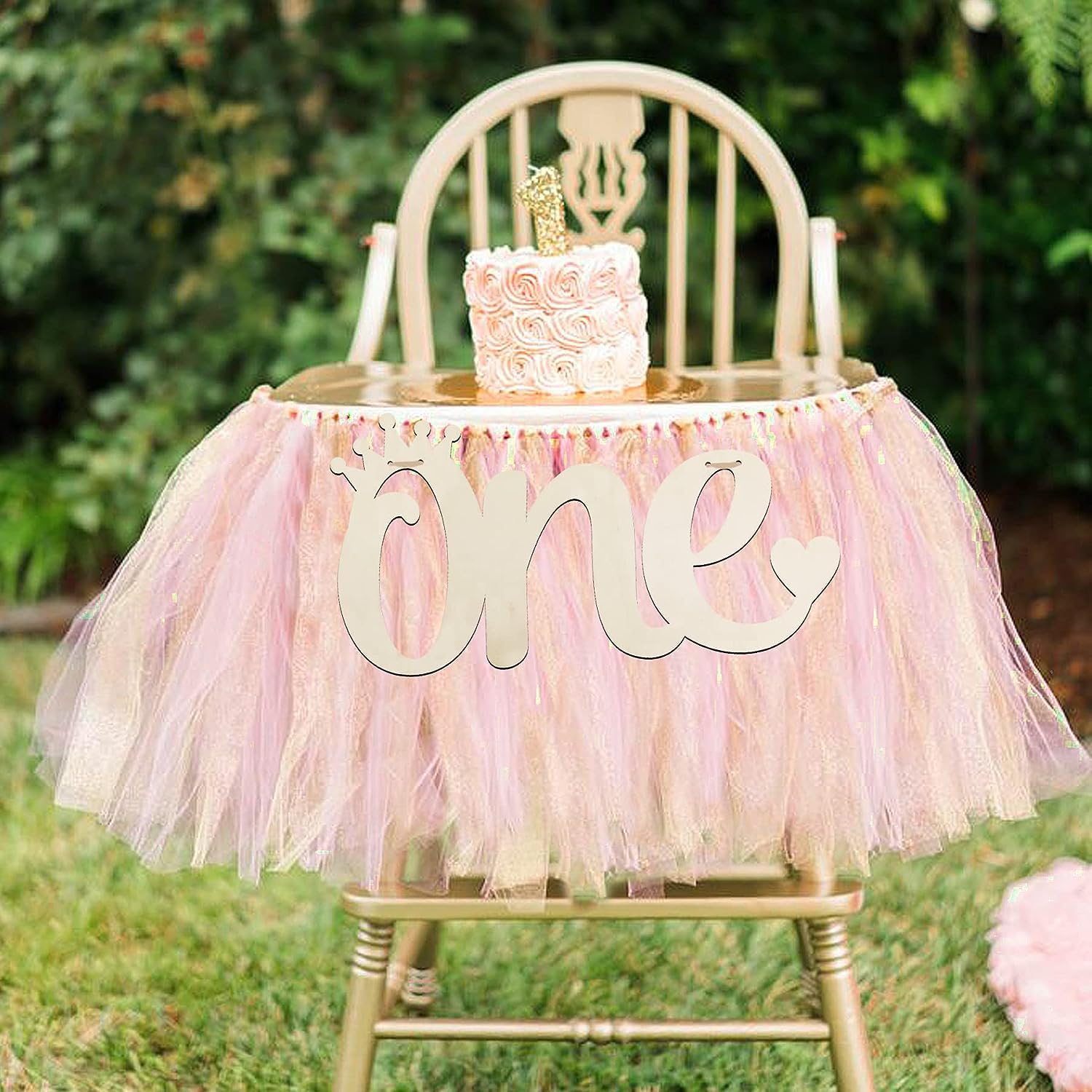 High chair with pink tulle skirt and 'one' sign, decorated with a small cake, outdoors.