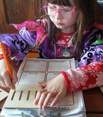 Child wearing glasses and a colorful jacket, engaged in writing or drawing on a piece of paper.