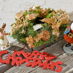 Decorative Christmas wreath with gold accents on a wooden surface, accompanied by festive figurines and 'Merry Christmas' sign.