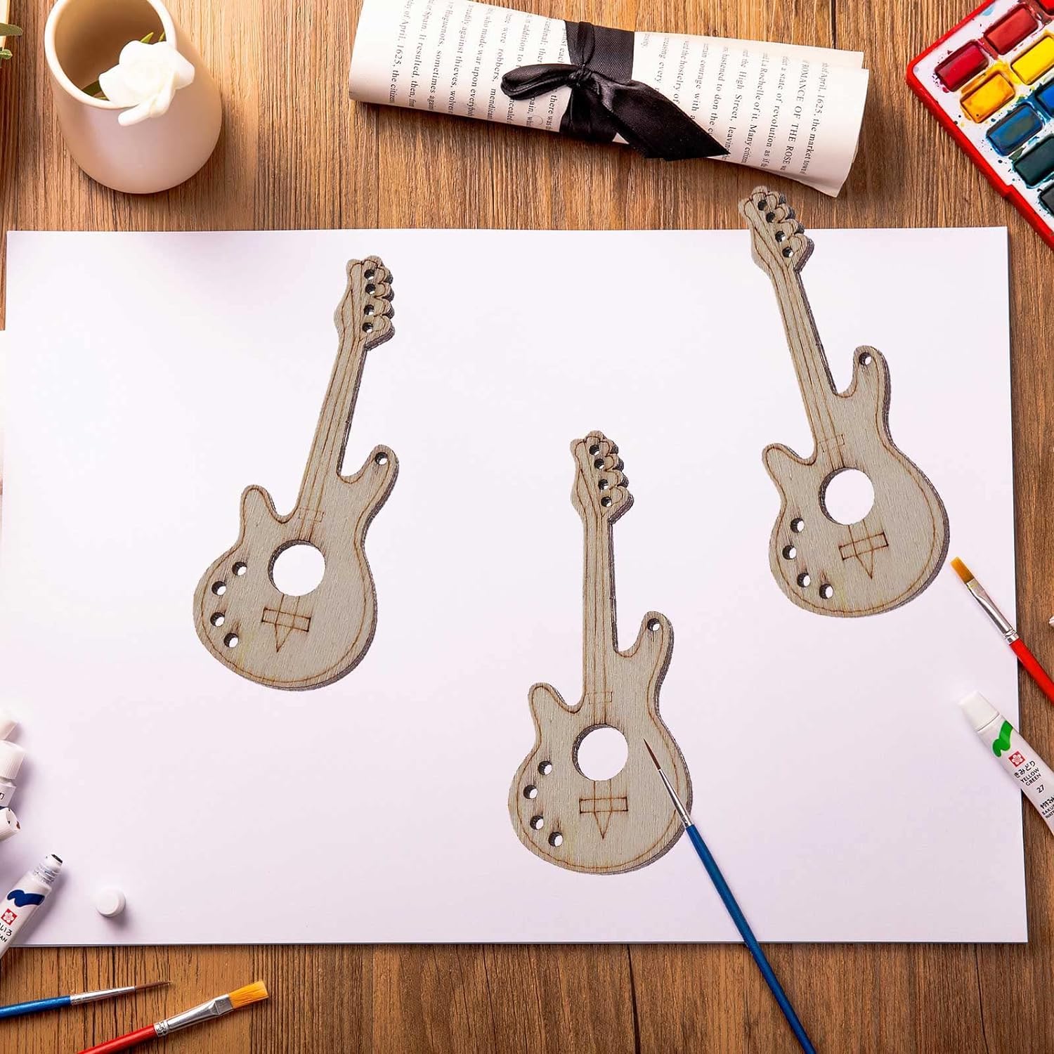 Three wooden guitar-shaped cutouts on a white sheet of paper with art supplies on a wooden table.