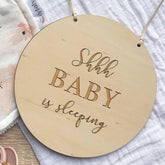 Round wooden sign with 'Shhh BABY is sleeping' text on a textured fabric background