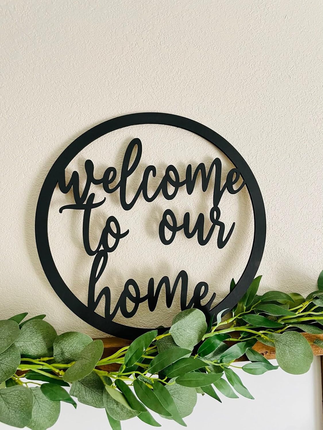Decorative wall art with 'welcome to our home' text and greenery on a neutral background