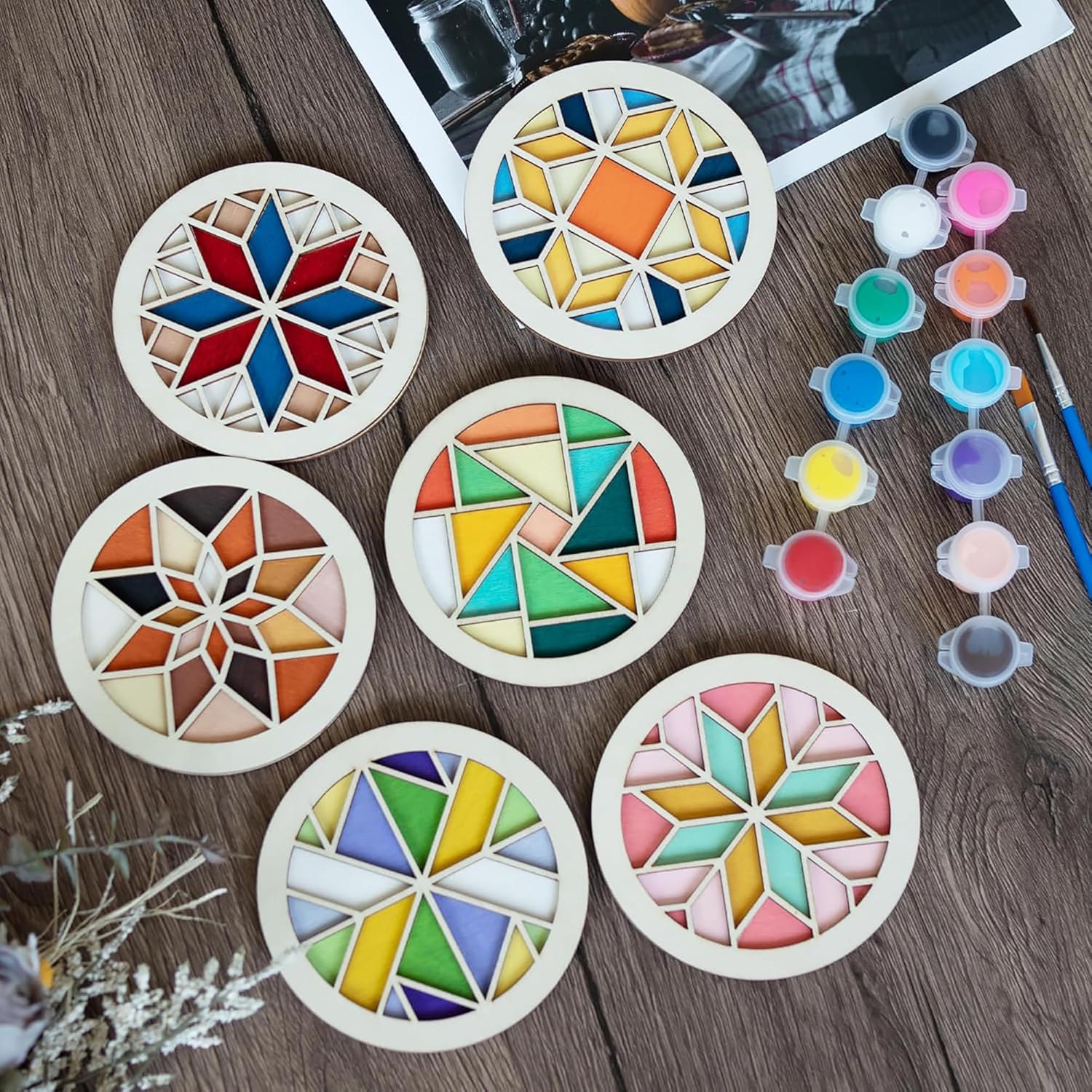 Set of colorful geometric coasters on a wooden surface with paint bottles and brushes.