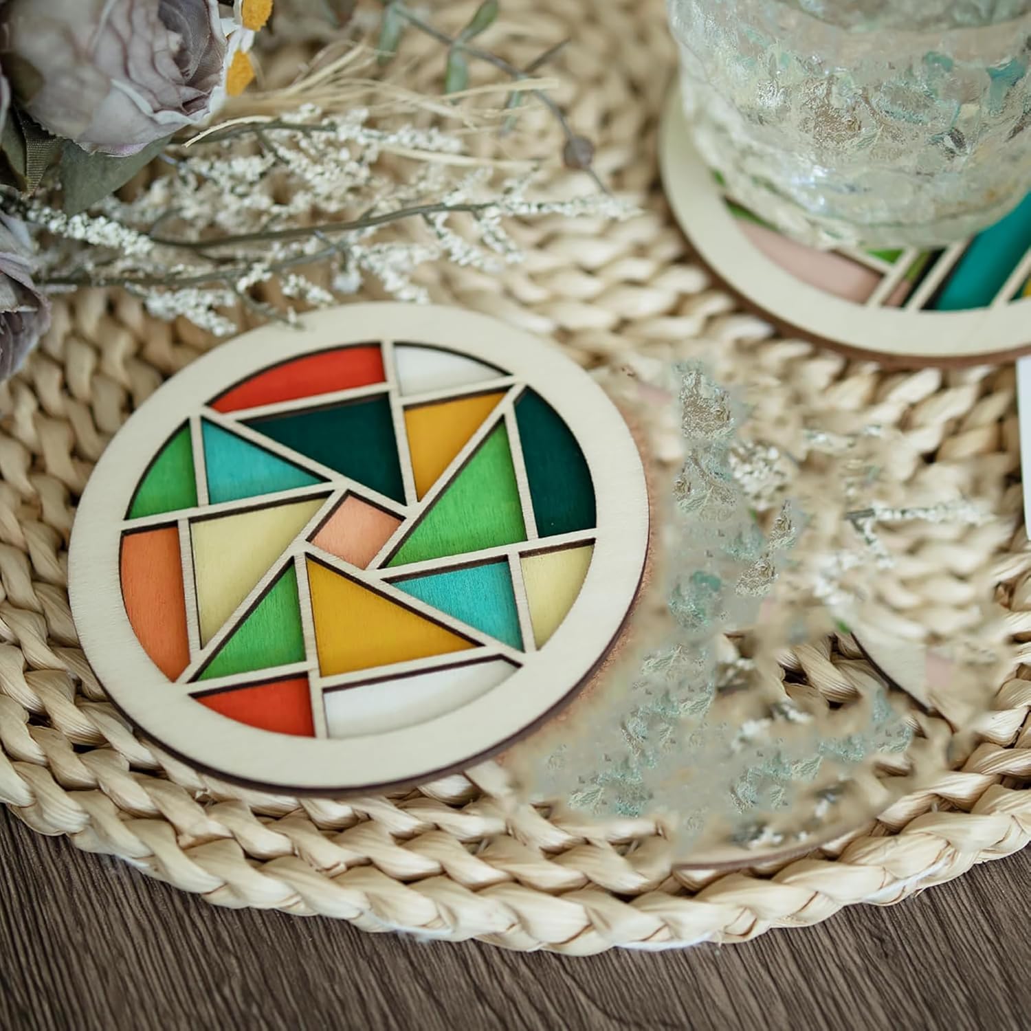 Colorful geometric coaster on a woven surface with flowers and a glass.
