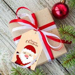 Gift wrapped in brown paper with a red ribbon and Santa Claus tag on a wooden surface with Christmas decorations.
