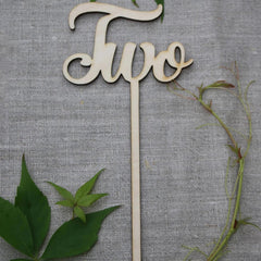 Wooden 'Two' cake topper with green leaves on a gray background