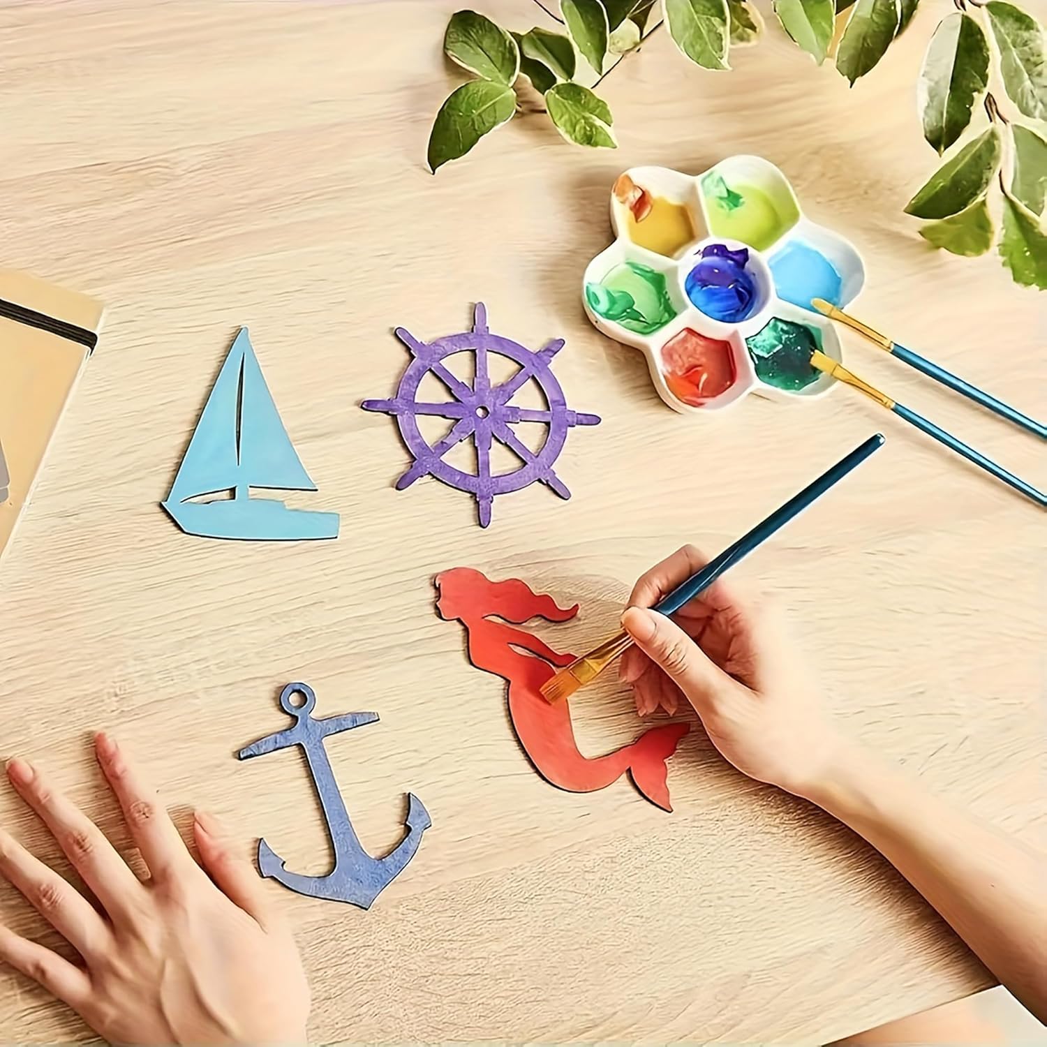 Colorful paper cutouts of a sailboat, wheel, and anchor on a wooden surface with a hand painting a mermaid figure.