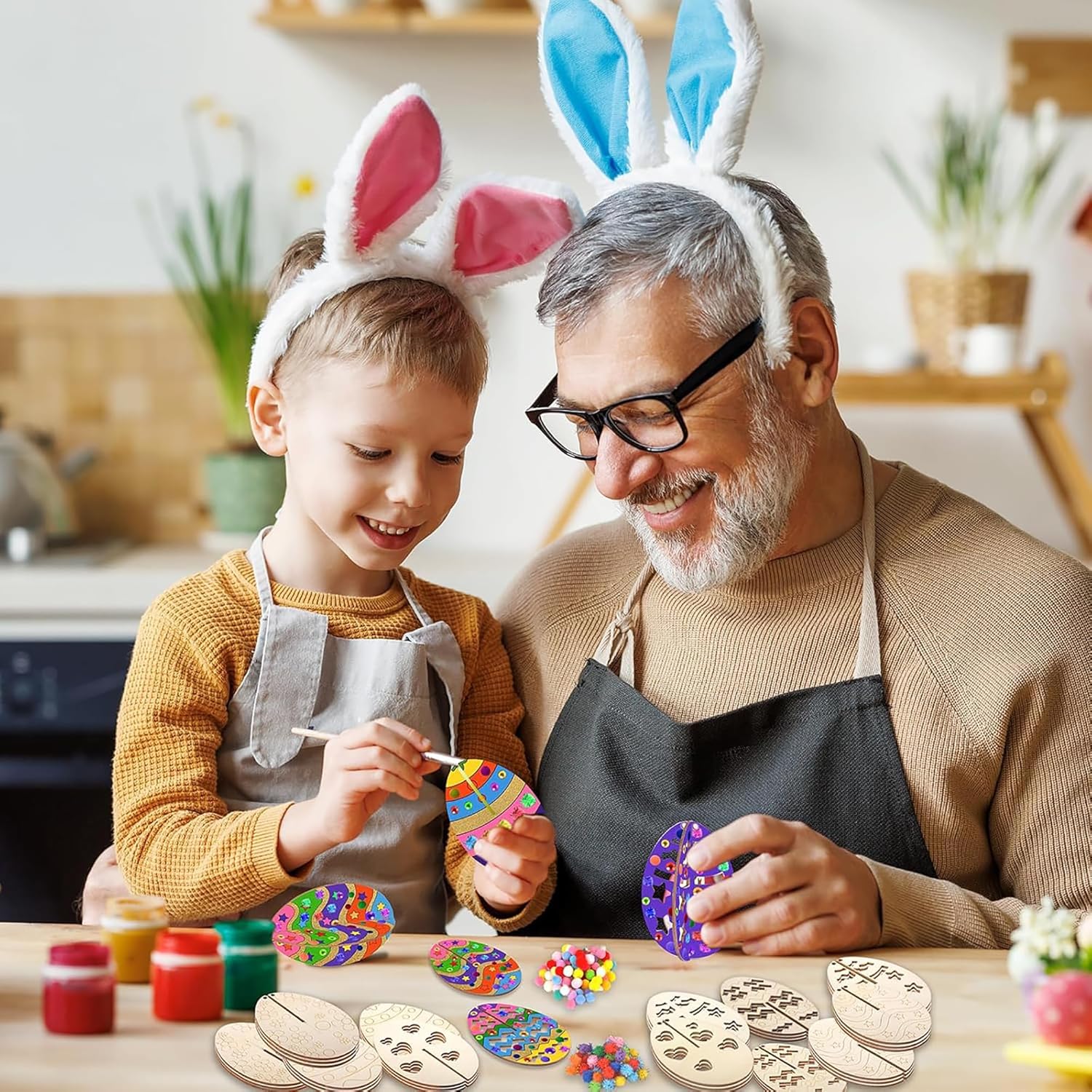 Man and child wearing bunny ears, painting wooden Easter eggs in a kitchen.