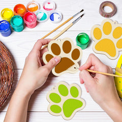 Hands painting wooden paw prints with colorful paint and brushes on a white surface.