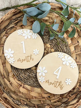 Two wooden milestone markers with floral designs and numbers on a woven mat.