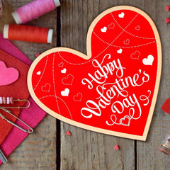 Heart-shaped wooden sign with 'Happy Valentine's Day' text on a wooden surface with sewing materials.