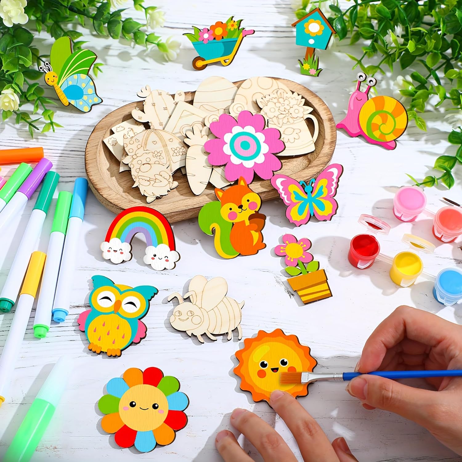 Children's craft materials including wooden shapes, markers, and paint on a table with greenery.