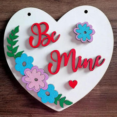 Heart-shaped decorative sign with 'Be Mine' text and colorful flowers on a wooden background