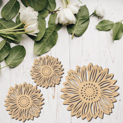 Three sunflower-shaped wooden coasters on a white wooden surface with green leaves and white flowers.