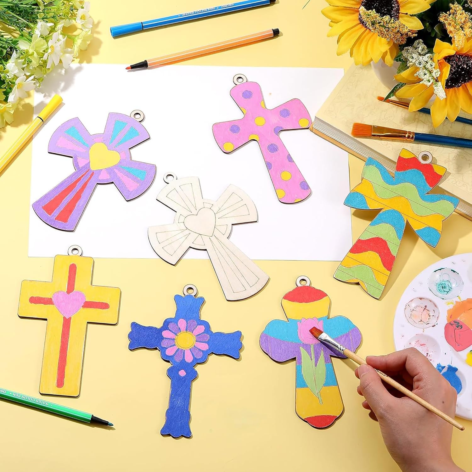 Colorful cross-shaped crafts on a yellow surface with painting tools and sunflowers.