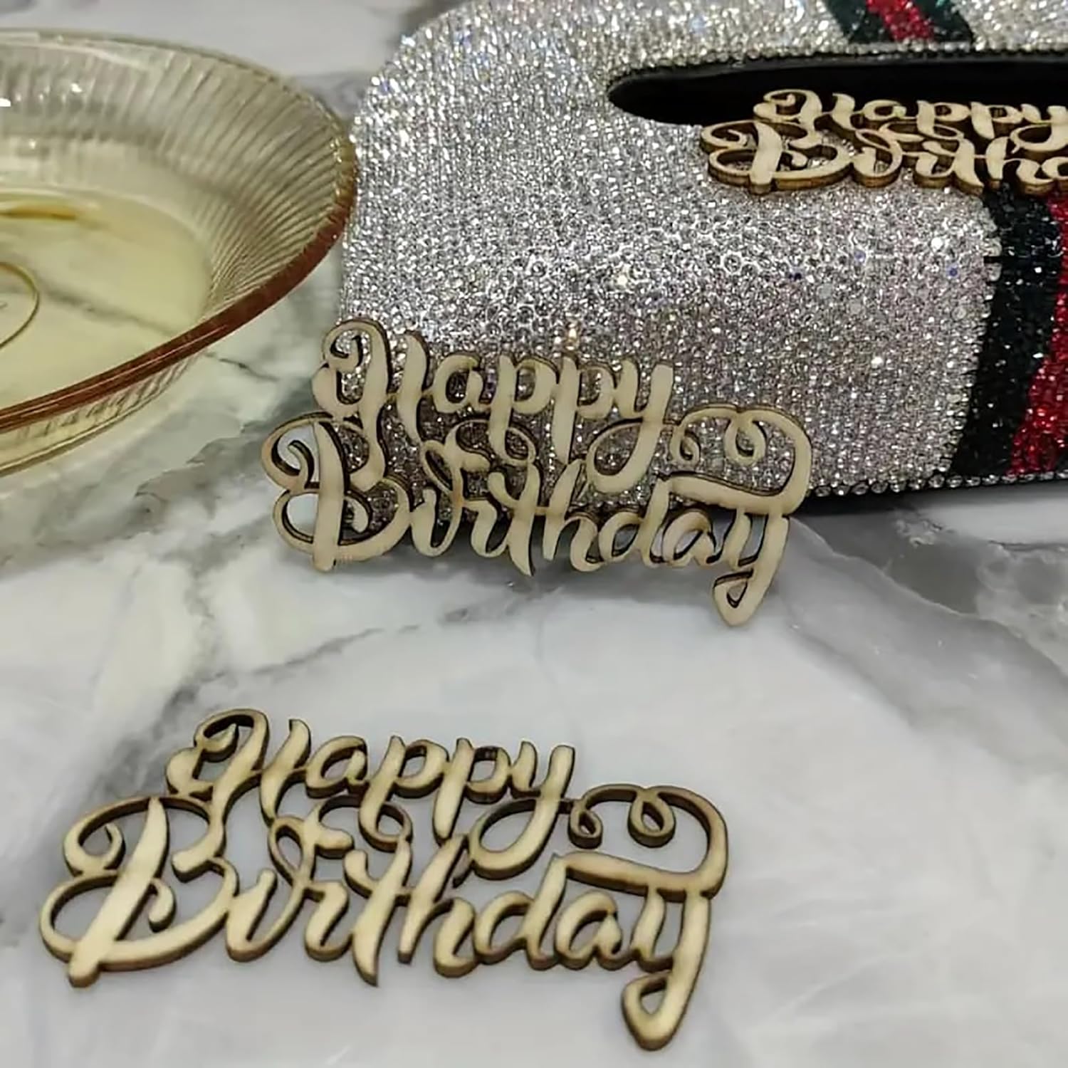 Wooden 'Happy Birthday' decorations on a marble surface with a sparkling decorative box in the background.