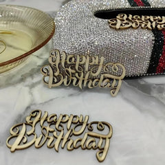Wooden 'Happy Birthday' decorations on a marble surface with a sparkling decorative box in the background.