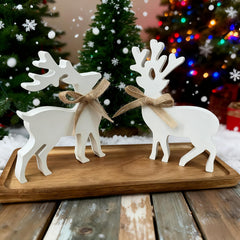 Two white deer figurines with brown bows on a wooden board in front of Christmas trees.