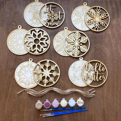 Decorative wooden ornaments with snowflake and text designs on a wooden surface, accompanied by paint bottles and brushes.
