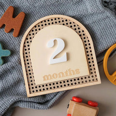 Wooden plaque with '2 months' on a textured surface with toys around