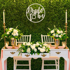 Decorated table with floral arrangements, candles, and 'Bride to be' sign against a green hedge background.