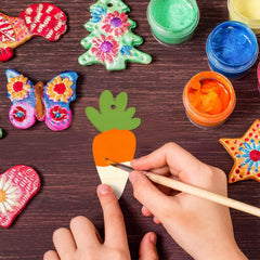 Colorful ceramic decorations on a wooden surface with hands painting a carrot-shaped piece.