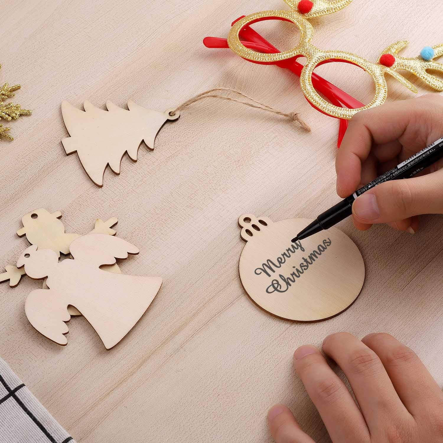 Hand writing 'Merry Christmas' on a wooden ornament with other ornaments and glasses in the background.