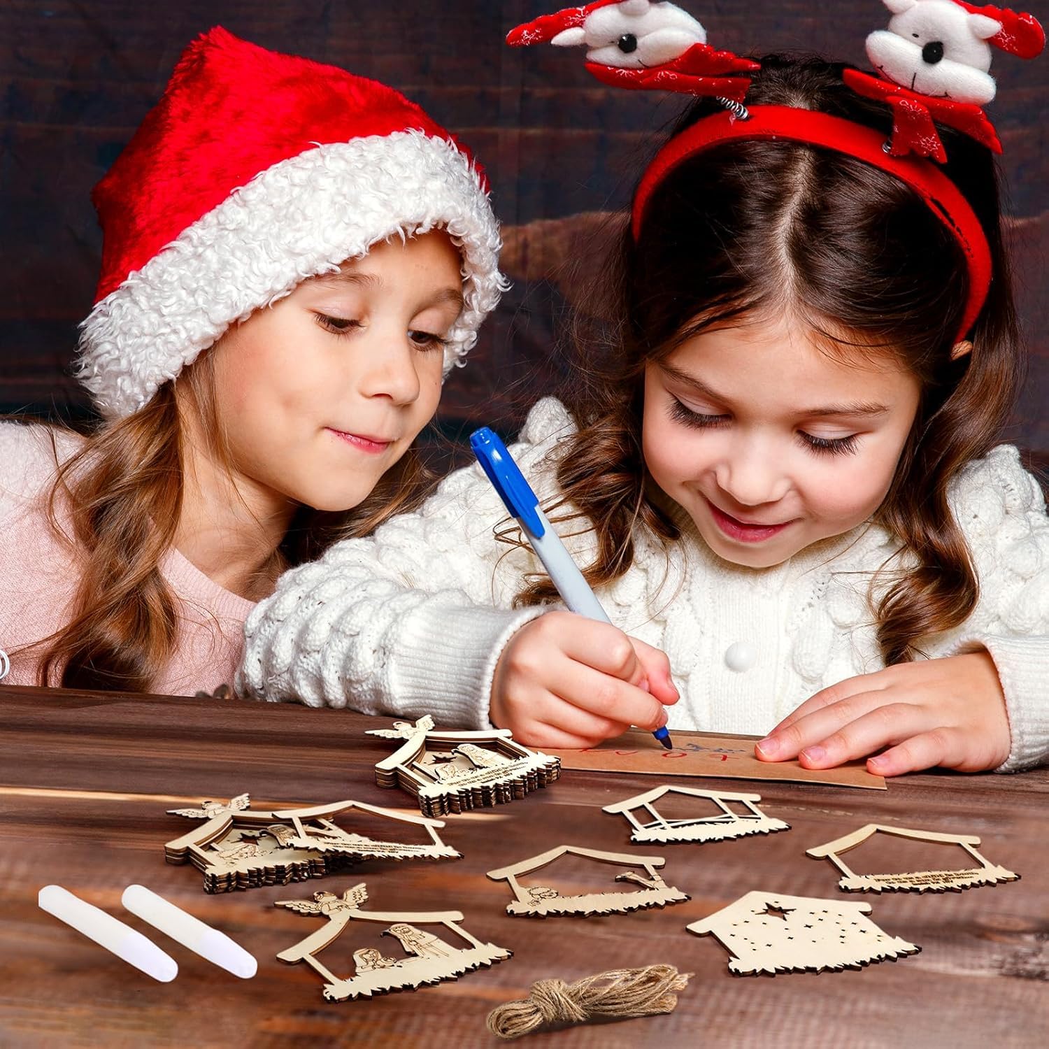 Two children wearing festive hats and reindeer ears, engaged in a craft activity on a wooden table.