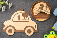 Wooden car-shaped milestone with number '1' and wooden plate with number '2', on a textured surface with flowers and a frog toy.