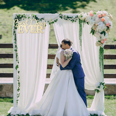 Couple embracing under a floral arch with 'Best Day Ever' sign on a sunny day.