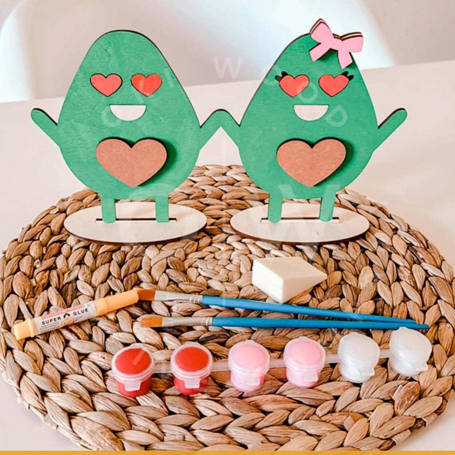 Two green wooden characters with hearts on a woven mat with paint supplies.