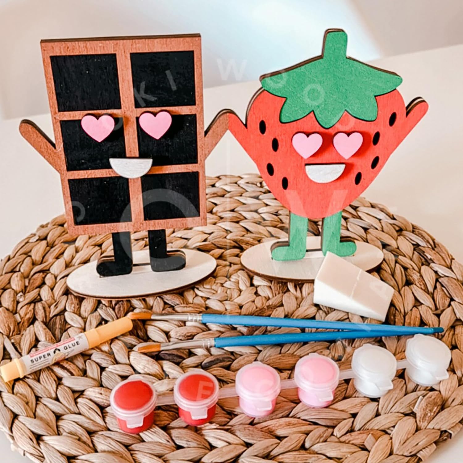 Two wooden figures, one resembling a house and the other a strawberry, with paint supplies on a woven mat.