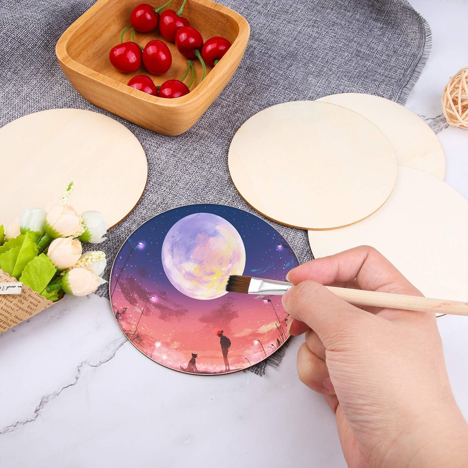 Person painting a colorful design on a round ceramic product with a brush, surrounded by ingredients on a marble surface.