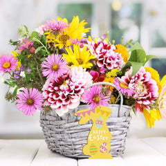 Bouquet of flowers in a basket with a 'Happy Easter' sign on a white surface.