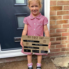 Young girl holding a 'My First Day' sign in front of a door.