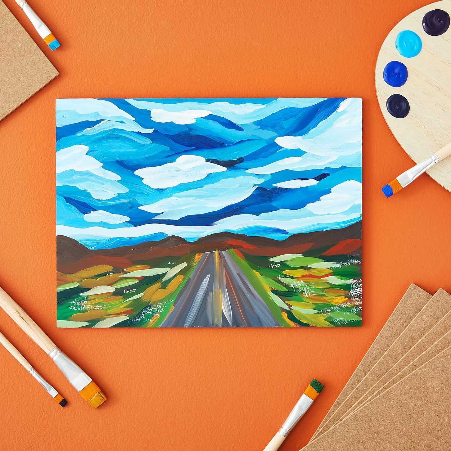 Painting of a road leading to mountains on an orange background with art supplies.