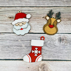 Decorative Christmas ornaments including a Santa Claus face, reindeer head, and red stocking with white snowflake design on a wooden background.