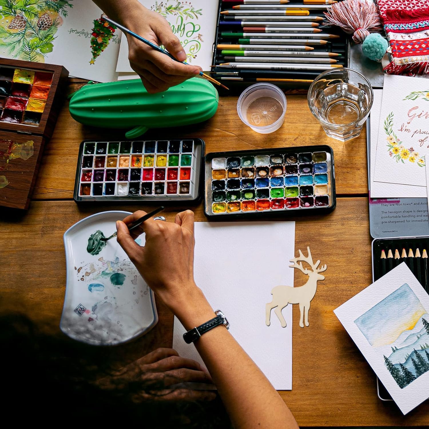 Artistic workspace with watercolor paints, brushes, and a person painting on paper.