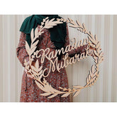 Decorative wreath with 'Ramadan Mubarak' text in front of a person wearing a green headscarf and patterned dress.