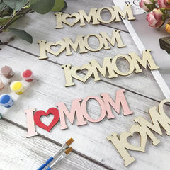 Wooden letters spelling 'I ❤️ MOM' on a wooden surface with paint bottles and brushes.