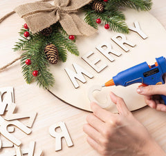 Hot glue gun being used to assemble a Christmas-themed wreath with wooden letters and decorative elements.
