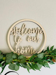 Wooden sign with 'welcome to our home' text surrounded by green foliage on a light background