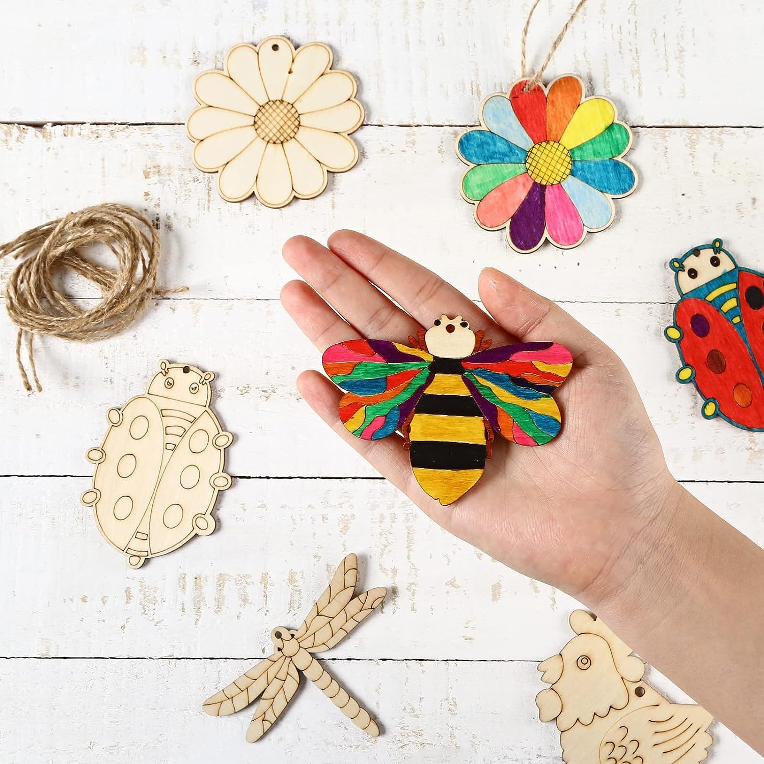 Hand holding a colorful bee ornament with wooden templates on a white wooden surface.