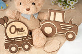 Wooden baby milestone card with teddy bear and tractor design on a soft surface.