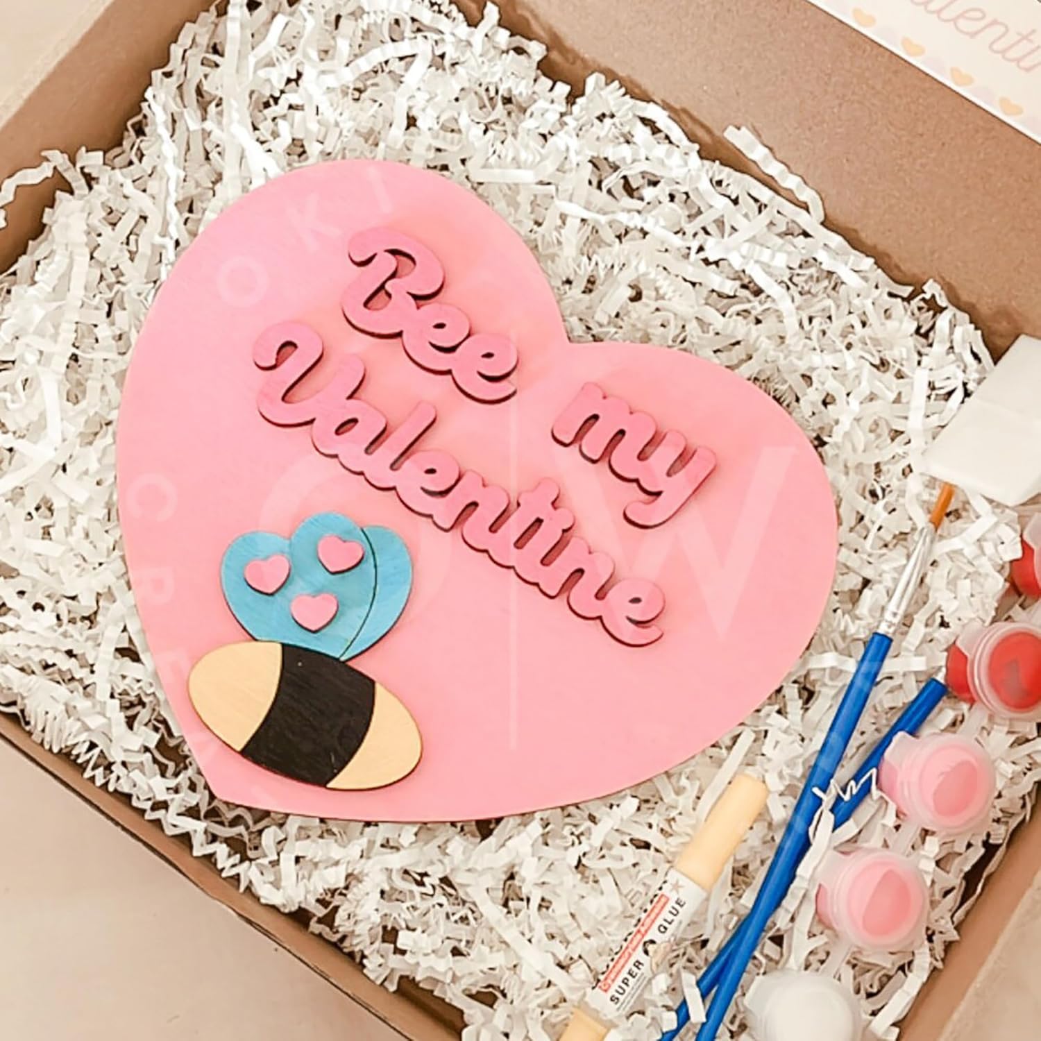 Pink heart-shaped card with 'Bee my Valentine' text, surrounded by paintbrushes and paint tubes in a box.